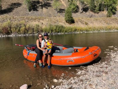 Family photo of mother, father and toddler on the Blackfoot River. They are seated on the side of a Zoo Town Surfers raft that is pulled up to shore and smiling as they enjoy a family adventure.