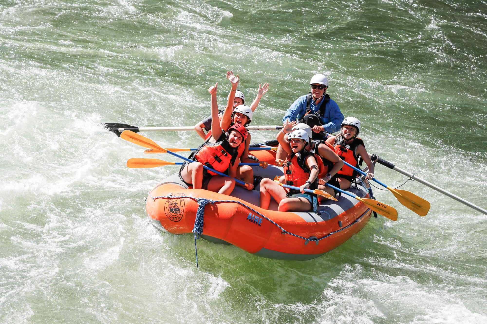 Group of happy rafters enjoying a sunny whitewater rafting trip on the Clark Fork River with Zoo Town Surfers. The rafters, in vibrant safety gear, cheer and smile while navigating the rapids, showcasing the fun and excitement of this outdoor adventure. The family is surrounded by exciting white water on the river!