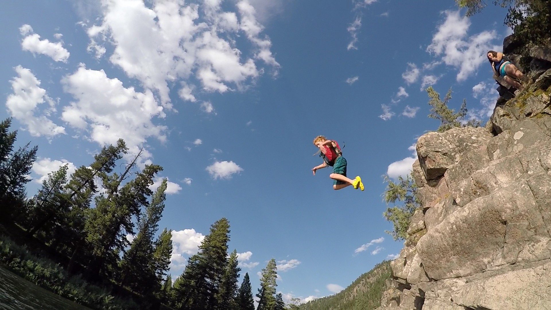 a kid jumping off a click on Blackfoot River. Blue sky with clouds and trees are in the background.